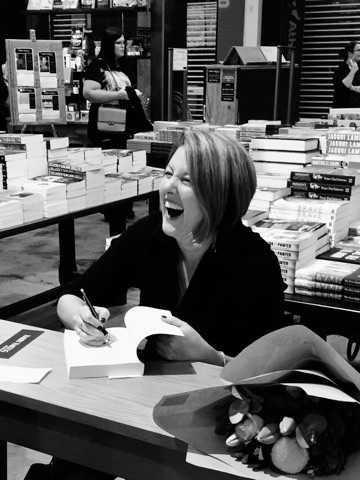 A woman sitting at a table. She is holding a pen and has paused in the act of writing something in the book open in front of her. She is laughing.