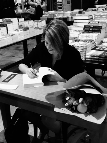 A woman sitting at a small desk with a book open in front of her. She is writing on the first page of the book. She is looking down at the book and smiling.